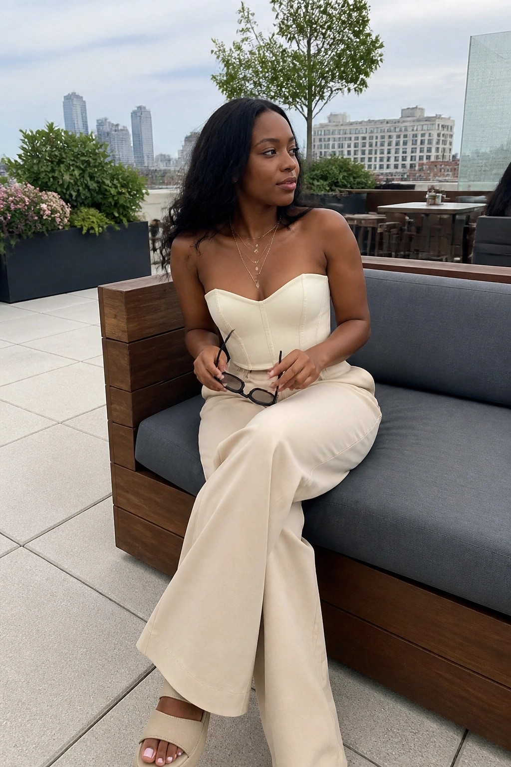 Woman in cream corset top and wide-leg pants on rooftop couch holding sunglasses