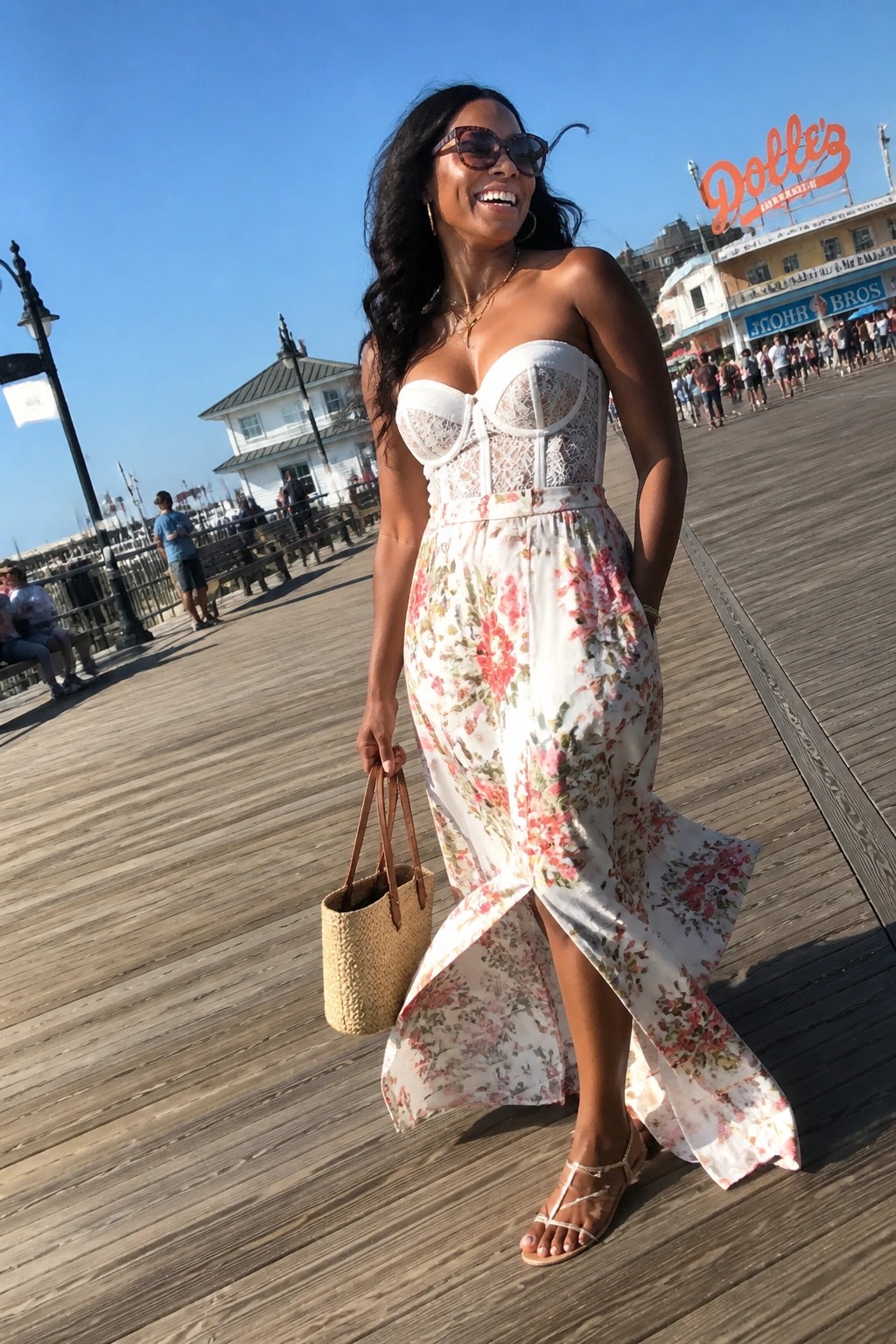 Smiling woman in white lace corset and floral maxi skirt on sunny boardwalk with straw bag
