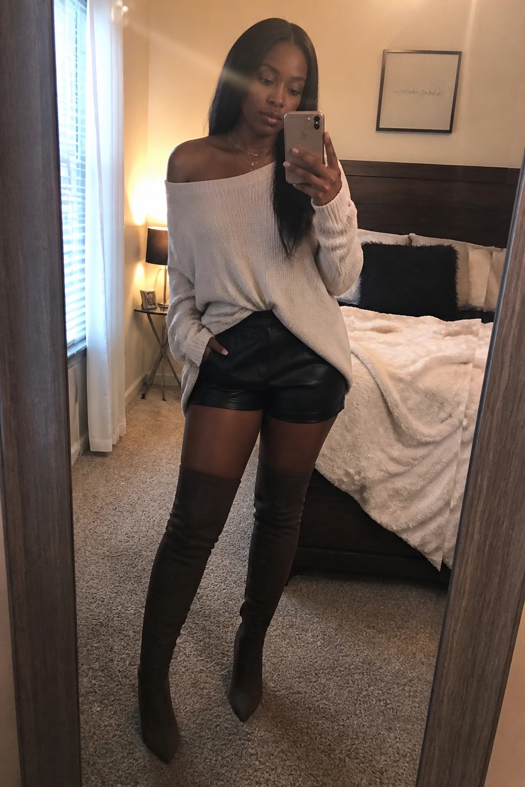 Woman in off-shoulder cream sweater and black shorts taking bedroom mirror selfie