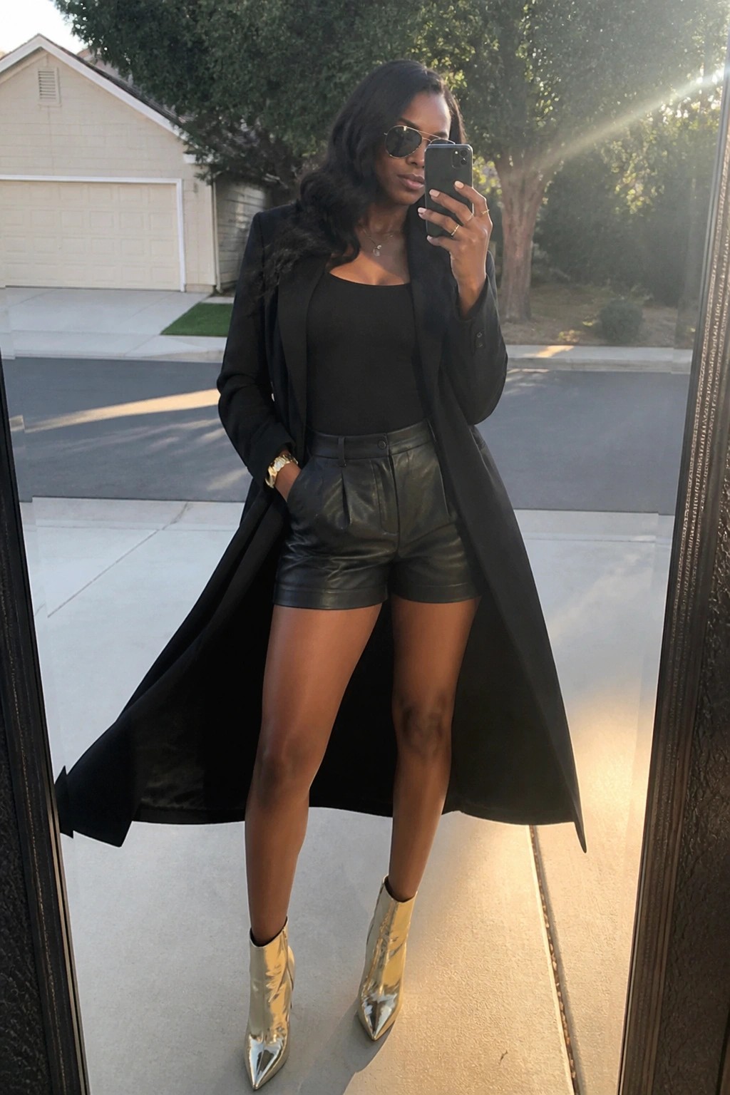 Woman in black coat and leather shorts takes selfie in gold boots outdoors