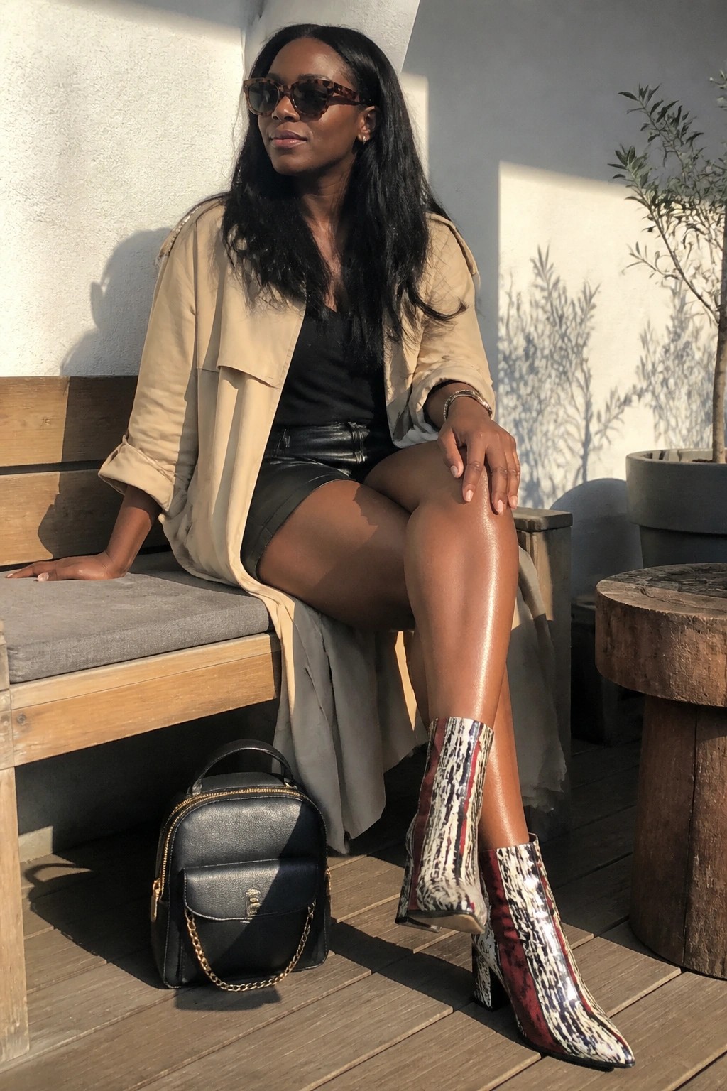 Woman in beige trench coat and snakeskin boots sits on bench beside black handbag.
