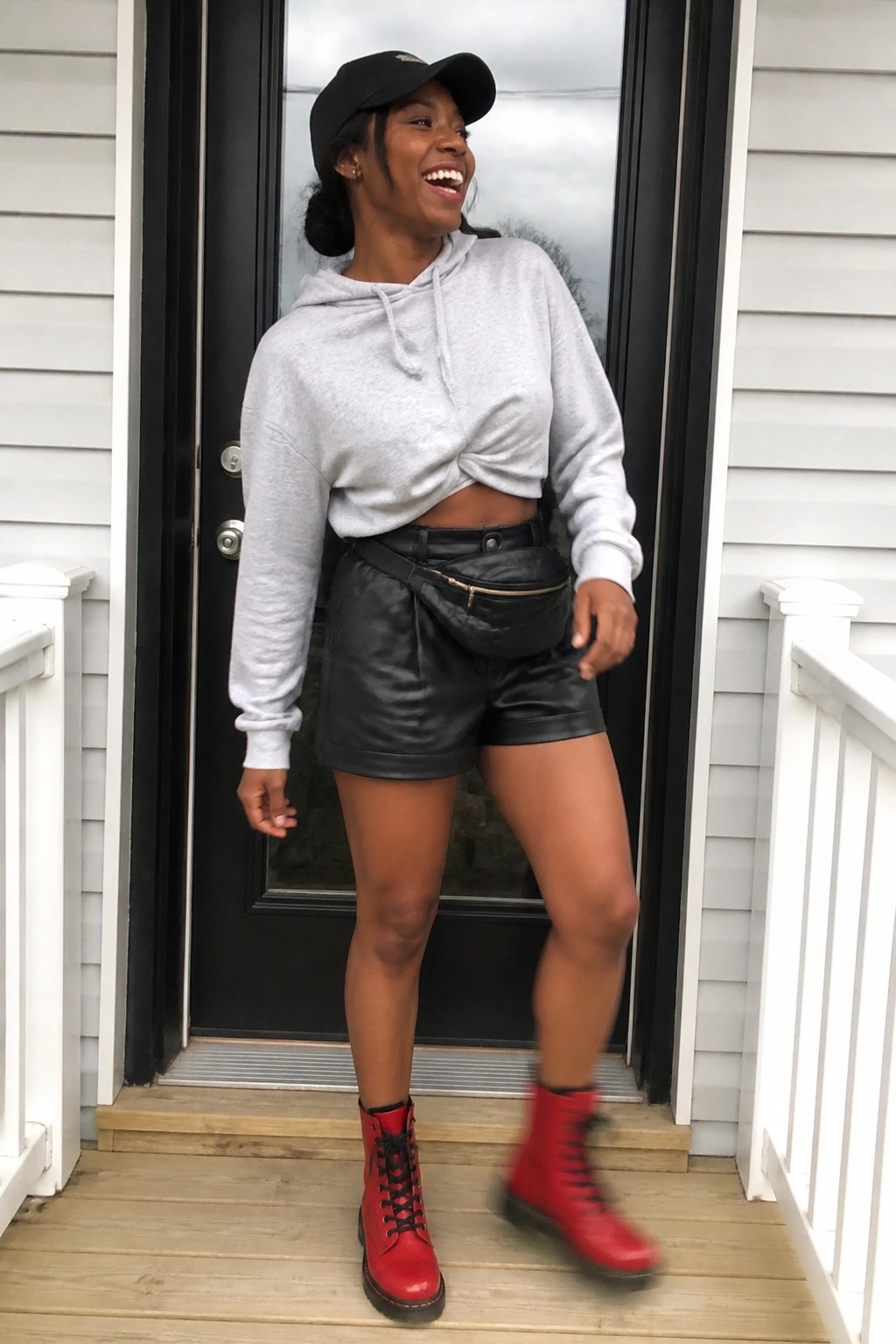 Smiling woman in black cap, cropped gray hoodie, leather shorts, and red boots on porch.