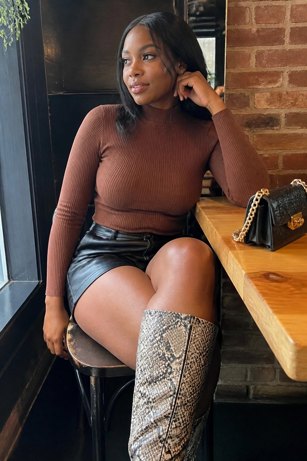 Woman in brown ribbed turtleneck tucked into black leather shorts paired with snake print boots and a black chain strap bag seated on a stool.