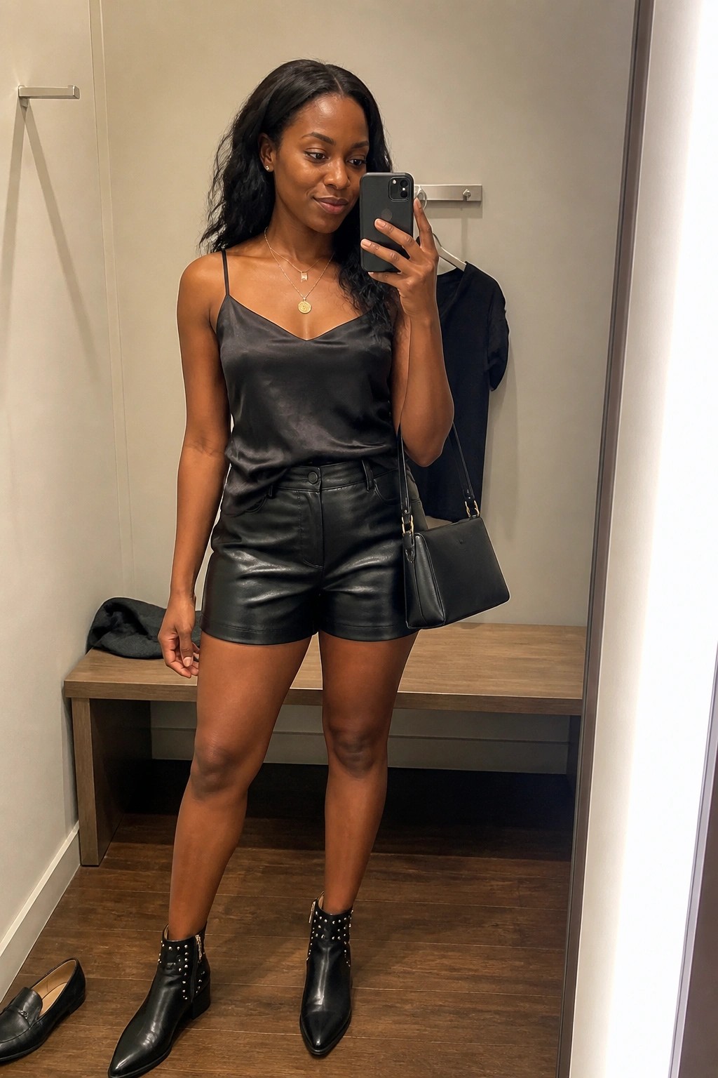 Woman in black satin camisole and leather shorts taking mirror selfie in fitting room