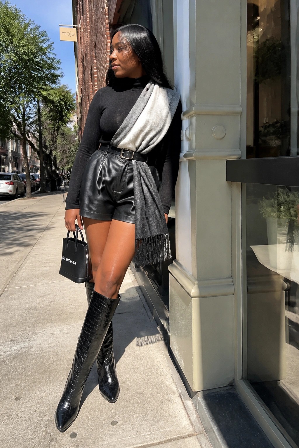 Woman in black turtleneck, leather shorts, scarf, and croc boots holding Balenciaga bag.