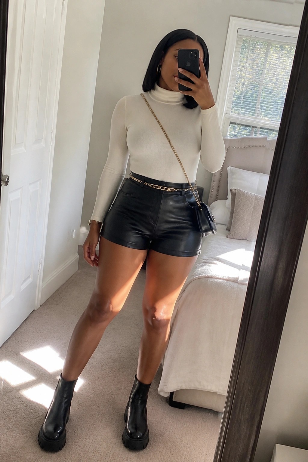 Woman in white turtleneck, black leather shorts, and boots taking mirror selfie in bedroom