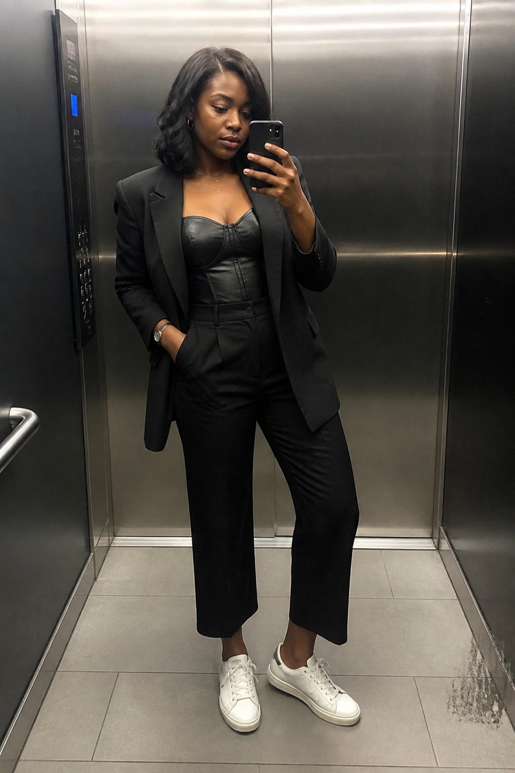 A Black woman stands in an elevator wearing a black leather corset top layered under an open black blazer with matching wide cropped trousers and white sneakers while holding a phone for a mirror selfie.