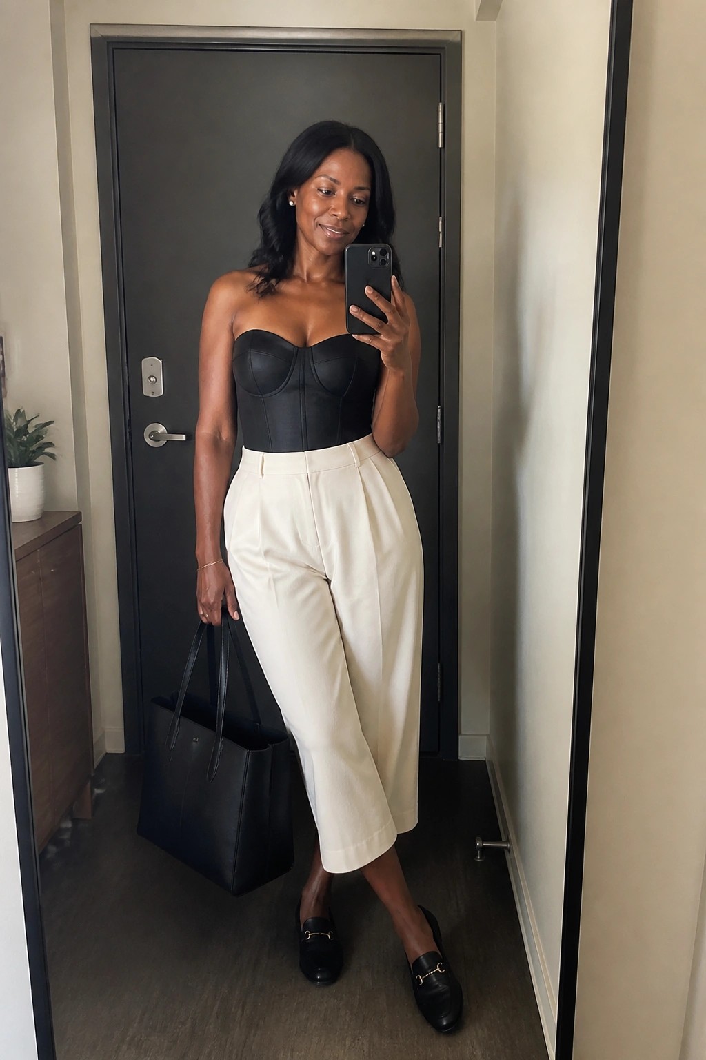 Woman in black strapless corset and white wide-leg pants taking mirror selfie with black bag
