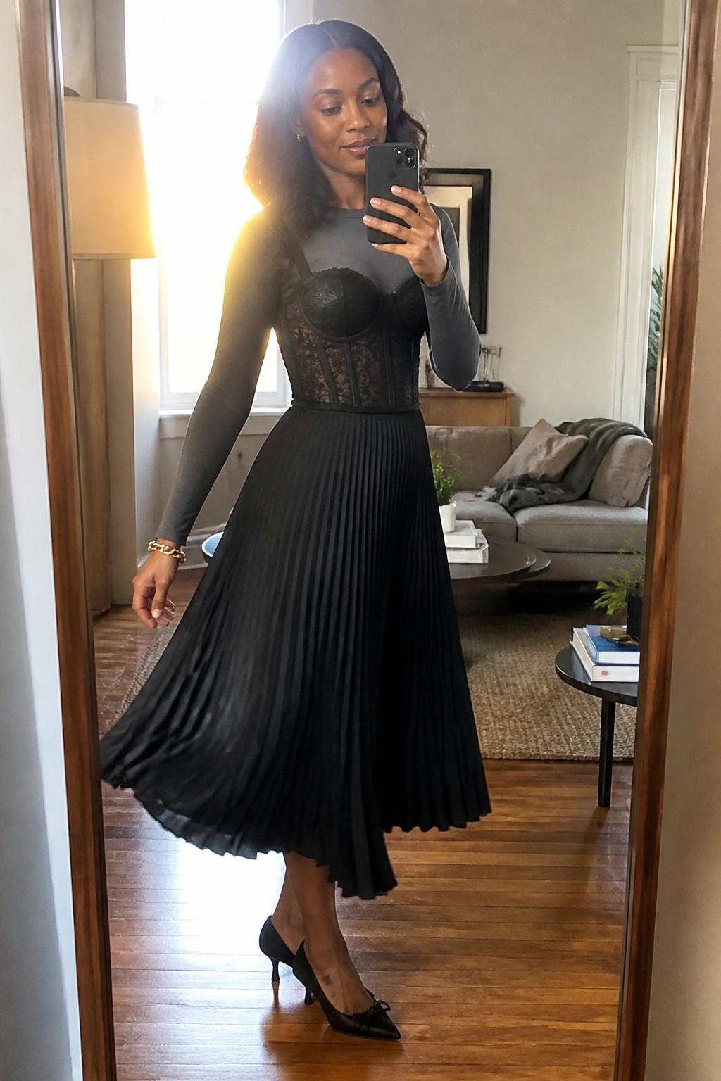 Woman wearing black lace corset over grey long sleeve top paired with black pleated midi skirt and black pointed heels with bow taking a mirror selfie