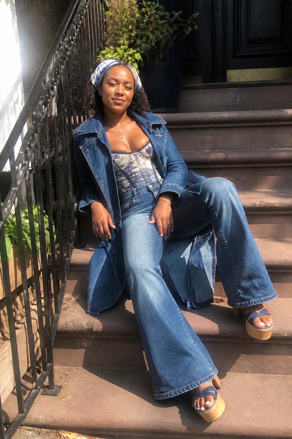 Woman in denim jacket, corset top, and flared jeans sits on stone steps wearing headscarf.