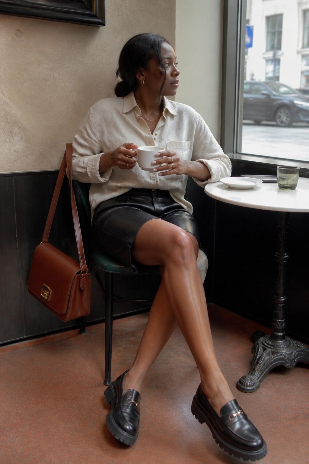 Woman in beige shirt and black leather skirt holding coffee by cafe window