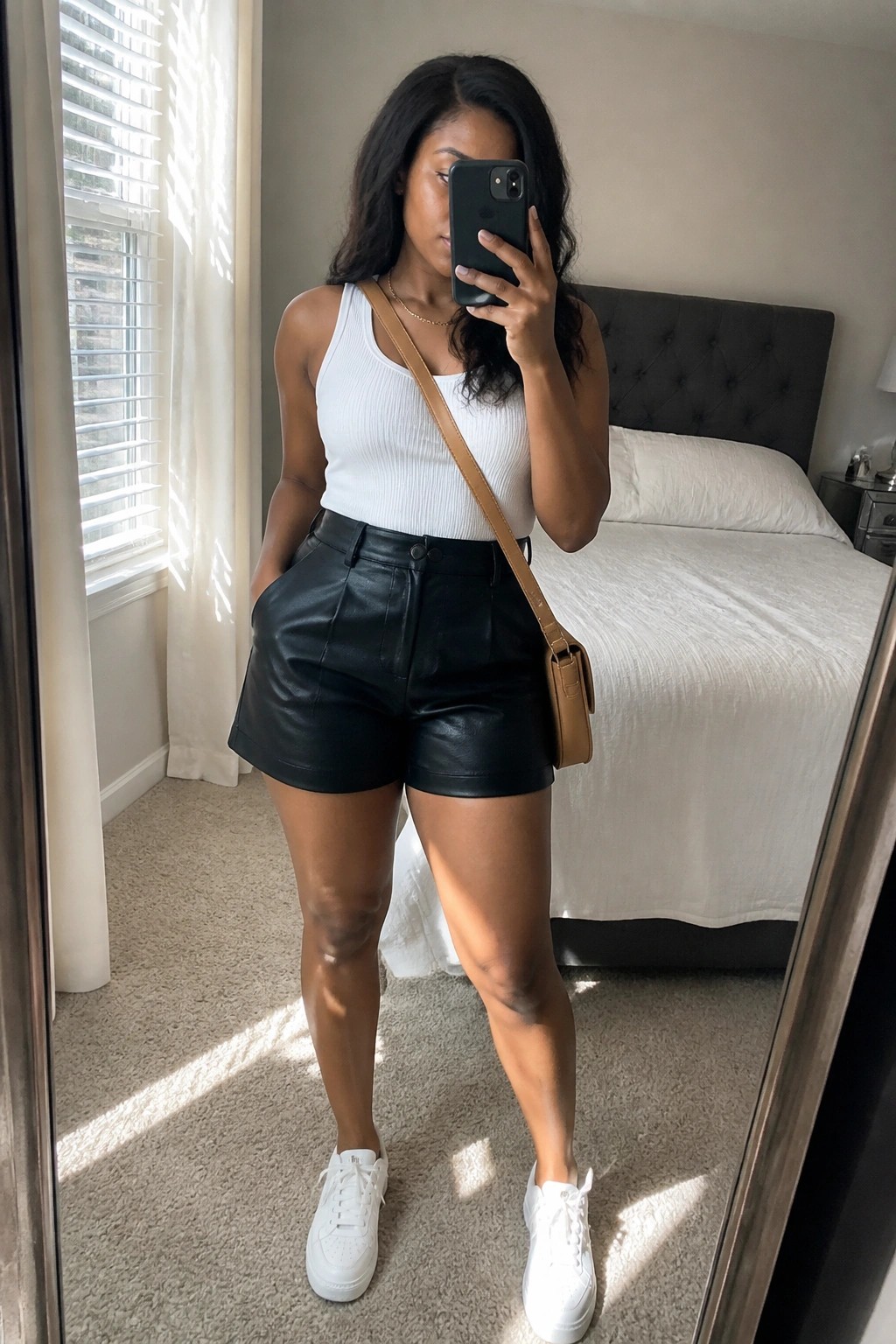 Woman in white tank top and black leather shorts taking mirror selfie in bedroom