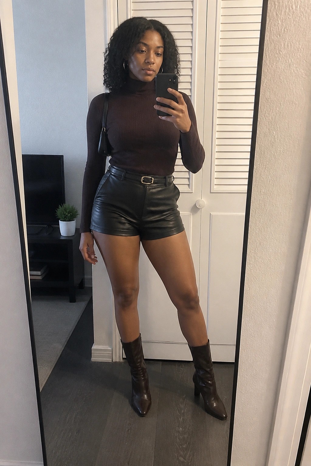 Woman in a brown ribbed turtleneck sweater tucked into black leather shorts with a black belt, paired with brown heeled ankle boots and a small black shoulder bag, taking a mirror selfie.