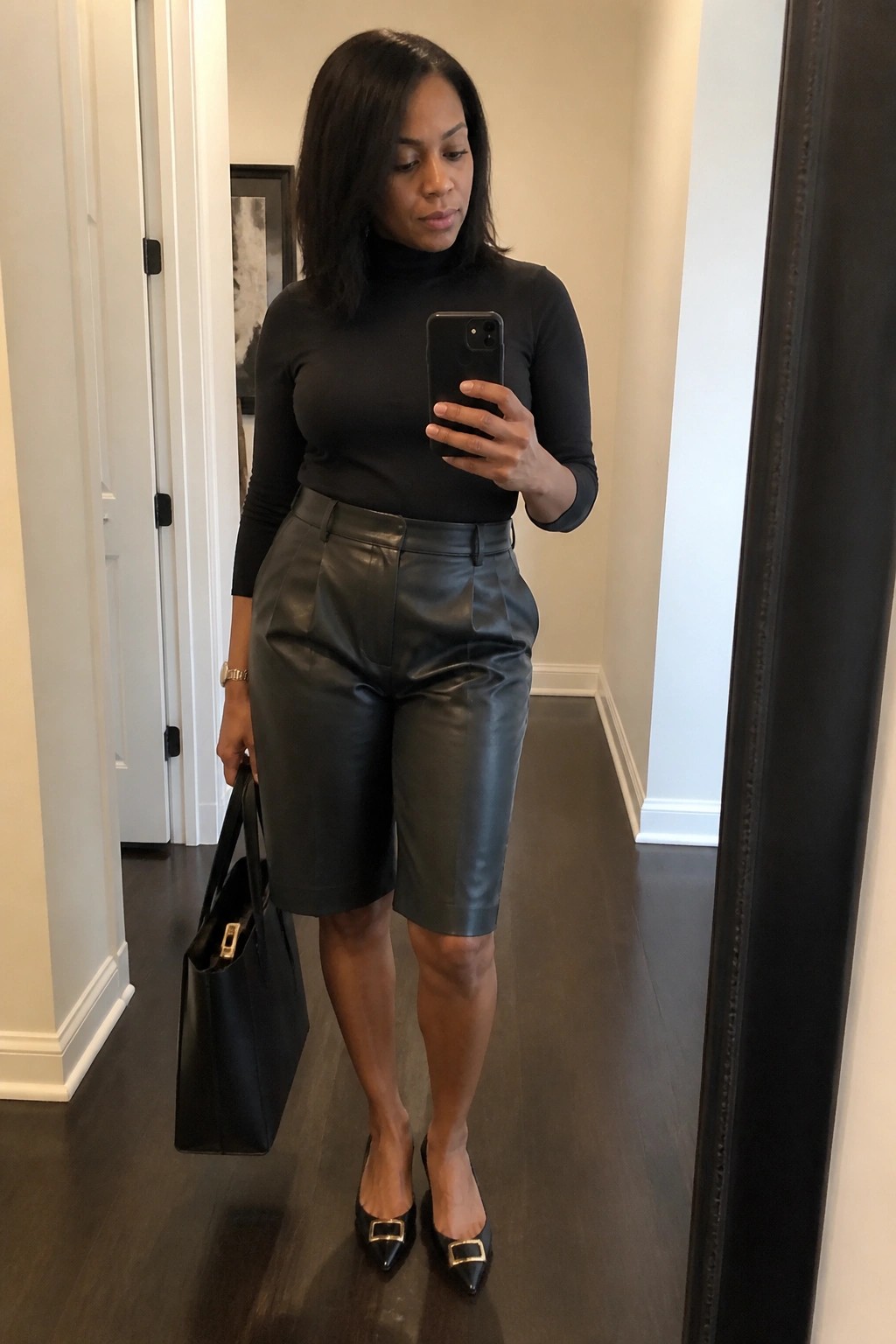 Woman wearing black turtleneck top tucked into black leather bermuda shorts with black pointed flats featuring gold buckles and carrying a black tote bag while holding a phone.