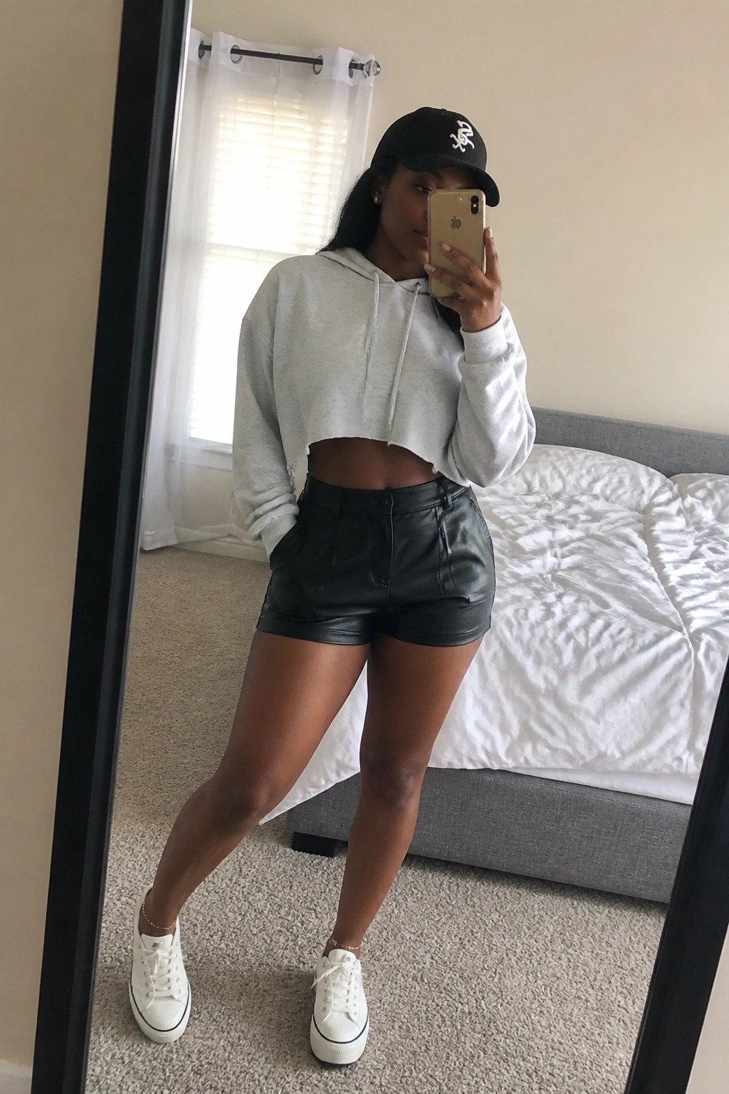 Woman in cropped gray hoodie and black shorts taking bedroom mirror selfie with phone
