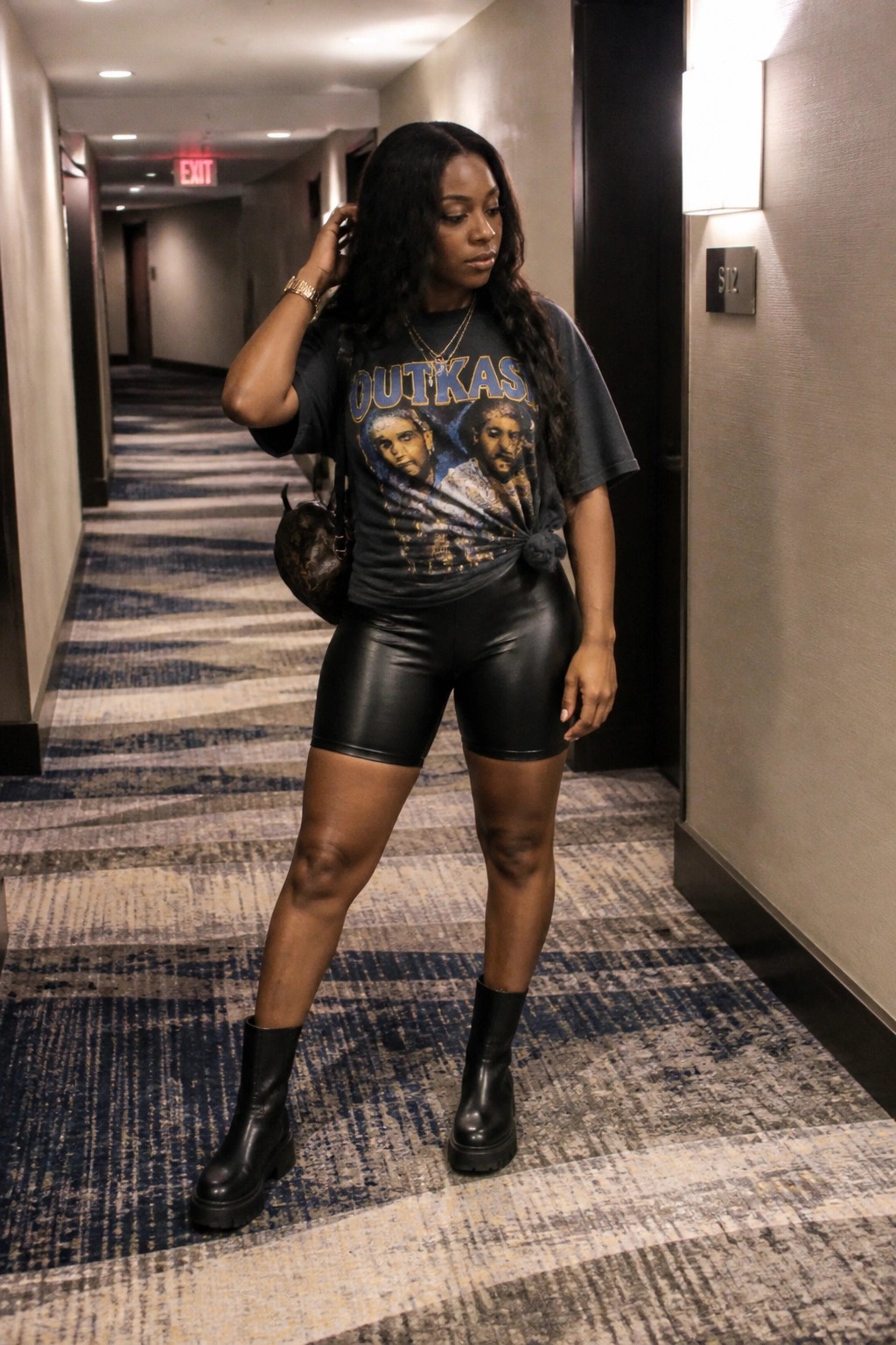 Woman in OutKast tee and black leather shorts posing in hotel hallway.