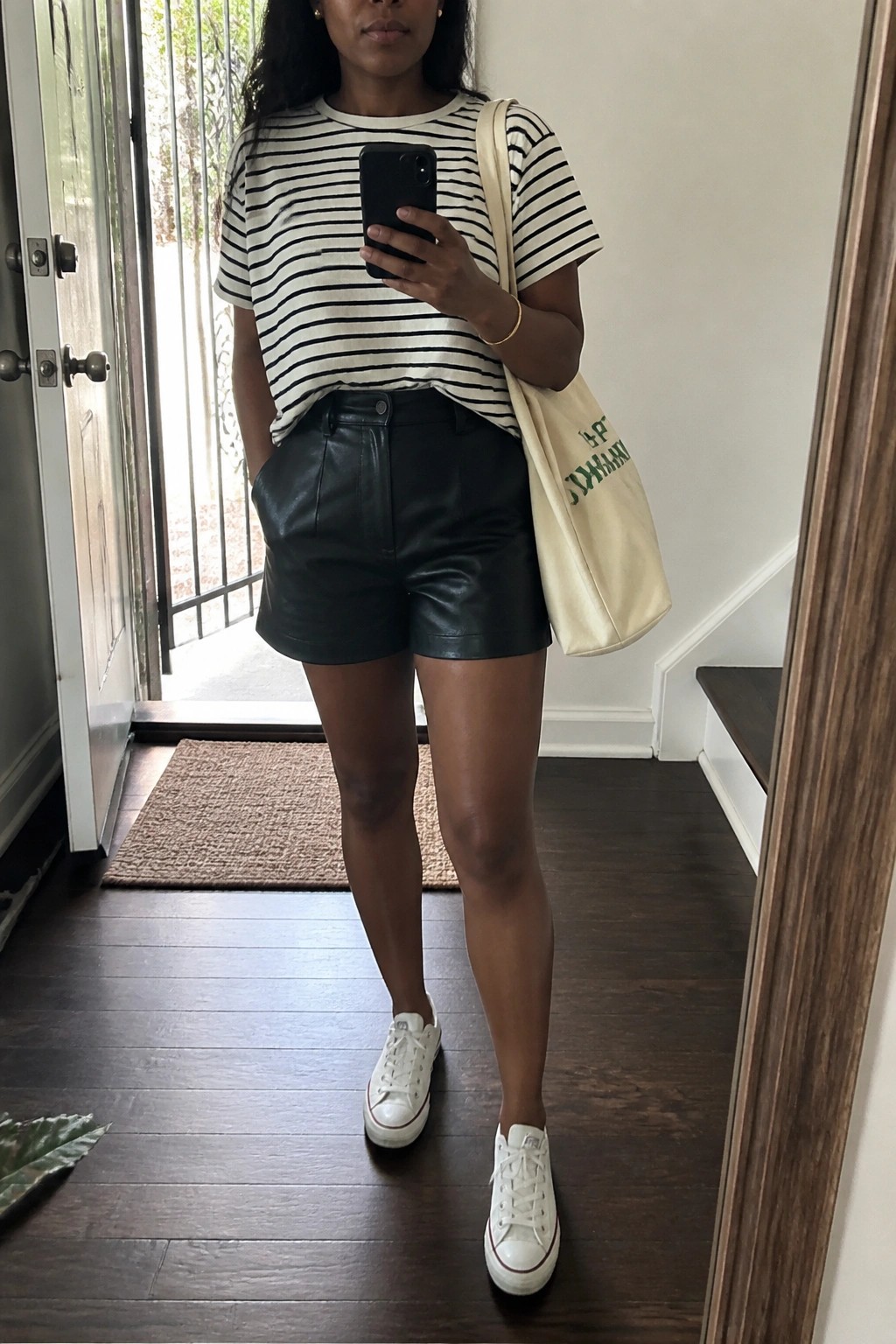 Woman in striped tee and black leather shorts taking mirror selfie with phone and tote bag