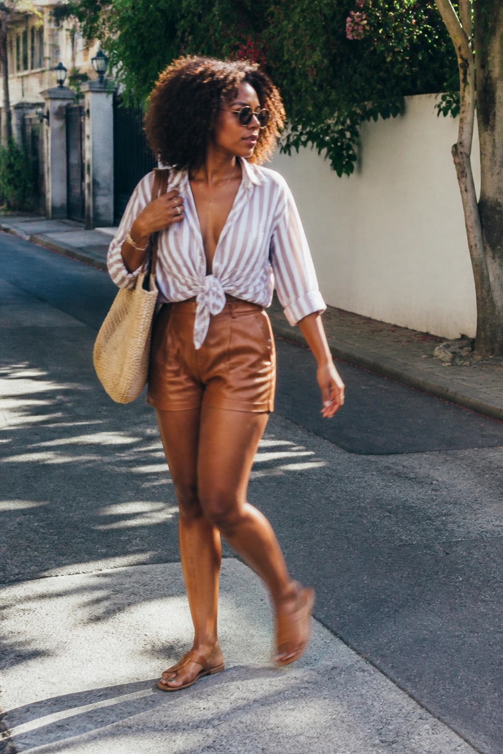 Woman with curly hair in tied striped shirt and brown shorts carrying woven bag on street