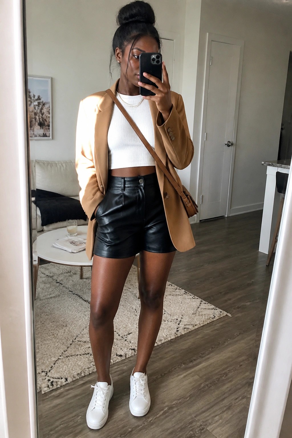 Woman in tan blazer, white crop top, black leather shorts taking mirror selfie in apartment