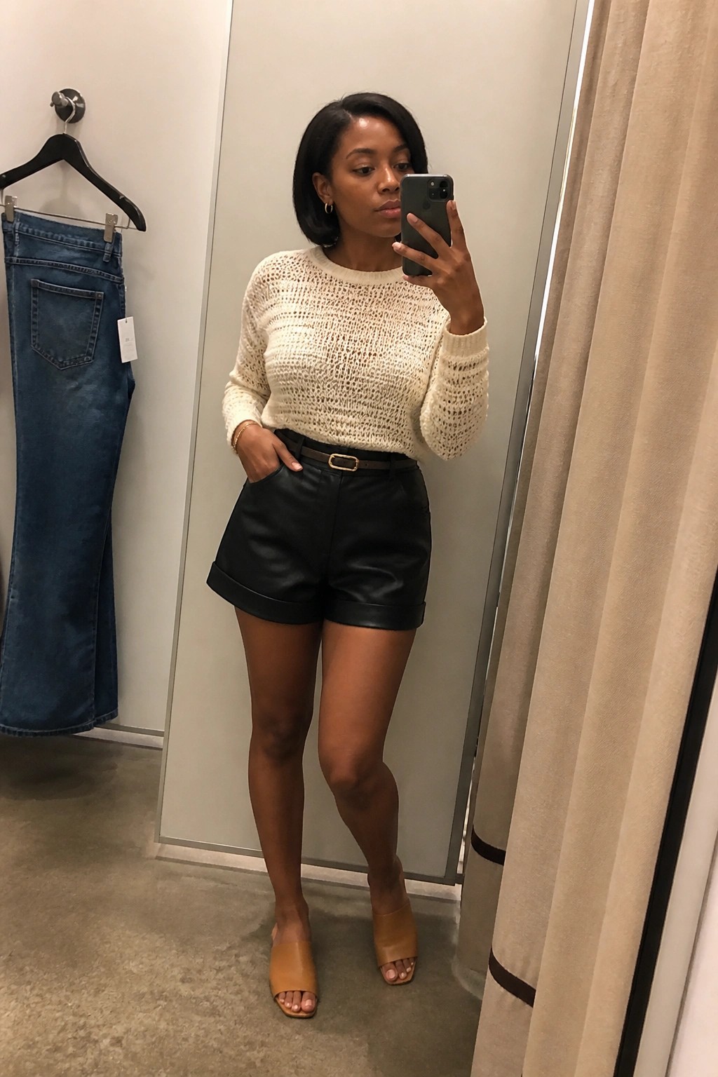 A Black woman taking a mirror selfie wearing black leather shorts with a brown belt, cream open knit sweater, and tan open toe slides.