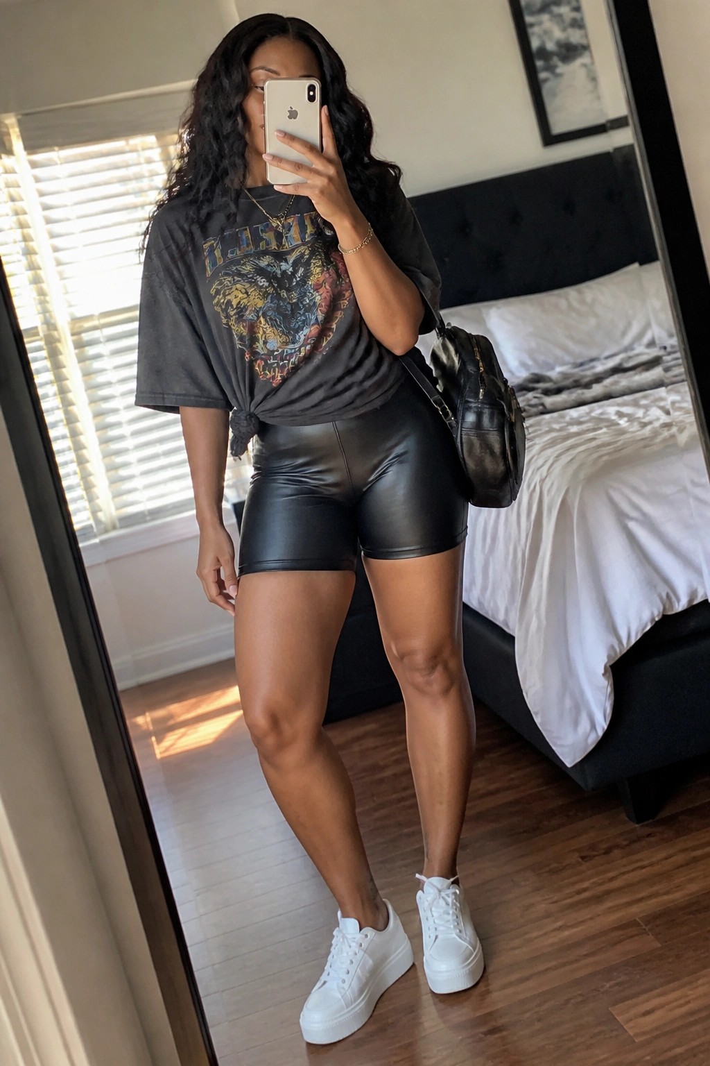 Woman in mirror selfie wearing black leather shorts, oversized dark graphic t-shirt tied at waist, white platform sneakers, black shoulder bag, and layered gold necklaces.
