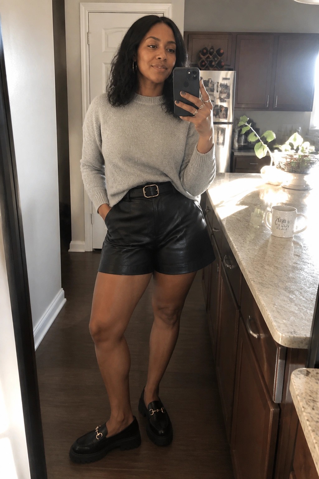 Woman in gray sweater and black leather shorts taking mirror selfie in kitchen