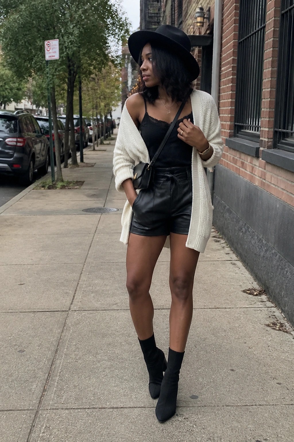 Woman in black hat, white cardigan, black top and shorts stands on city sidewalk.