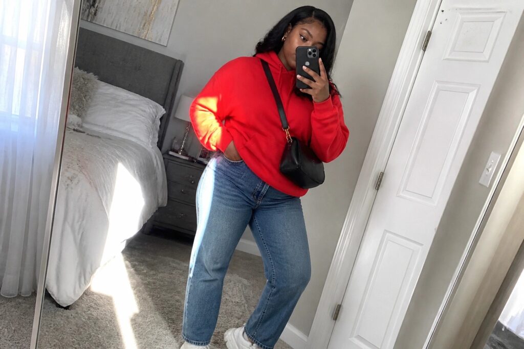 23 Cool Red Streetwear Outfits for Black Women Centered Around Casual Confidence Woman wearing an oversized red hoodie tucked slightly into straight-leg blue jeans with white sneakers and a small black crossbody bag taking a mirror selfie in a bedroom