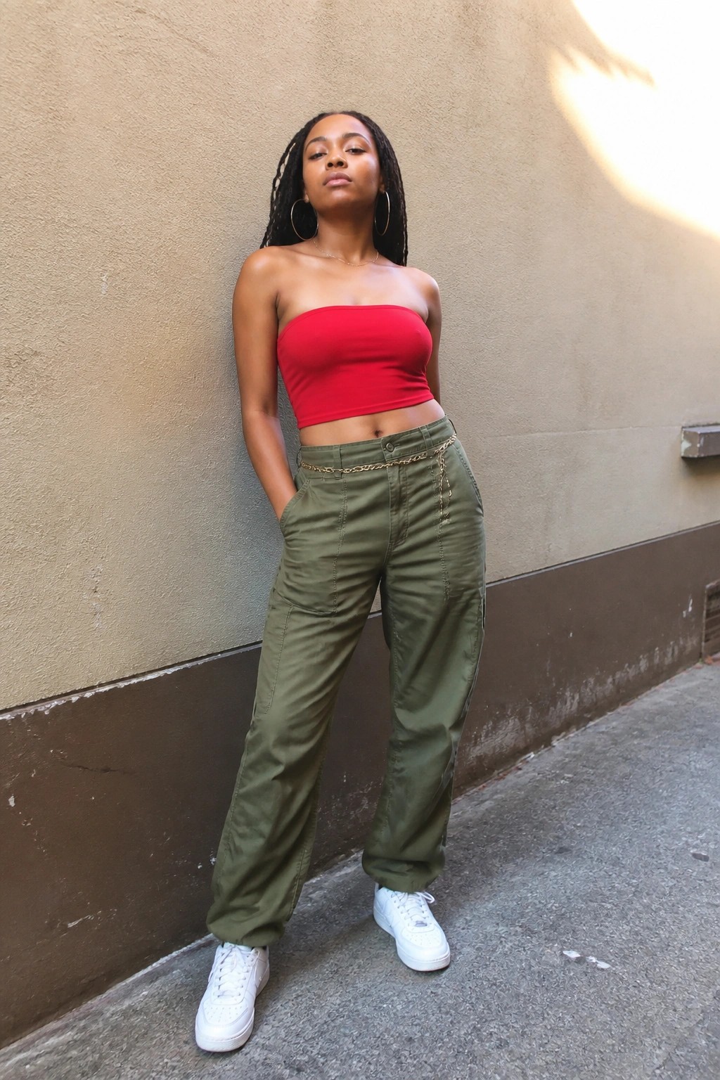 A Black woman wearing a red strapless crop top, olive green cargo pants with a gold chain belt, and white sneakers, standing against a wall.
