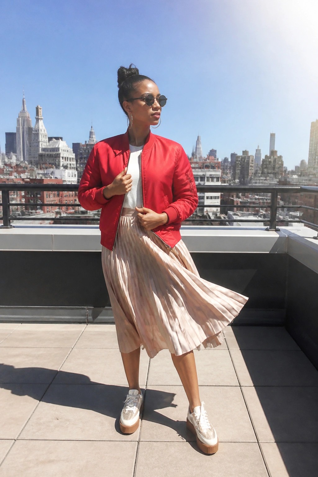 Woman in red bomber jacket and pleated pink skirt on sunny city rooftop with skyline