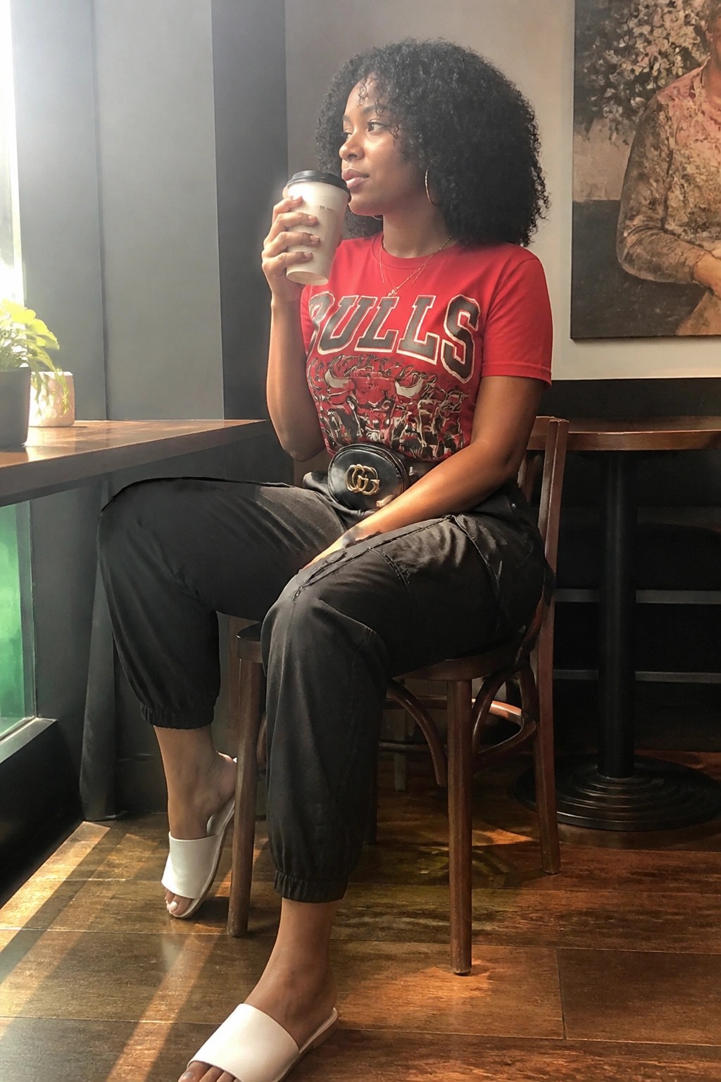 Woman in red Bulls shirt sips coffee at wooden cafe table wearing black pants.