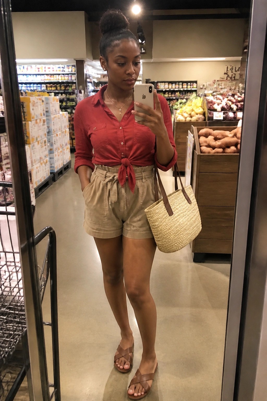 Woman in red tied shirt and beige shorts taking mirror selfie in grocery store.