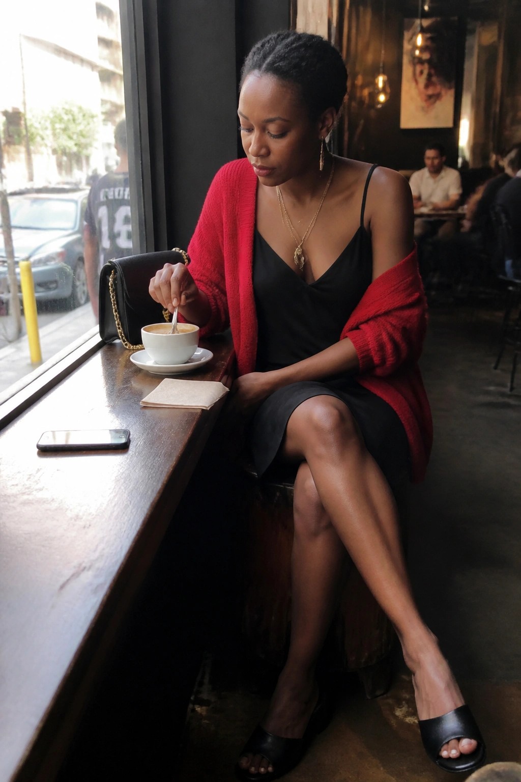 Woman in red cardigan stirs coffee at cafe table by window with black handbag.
