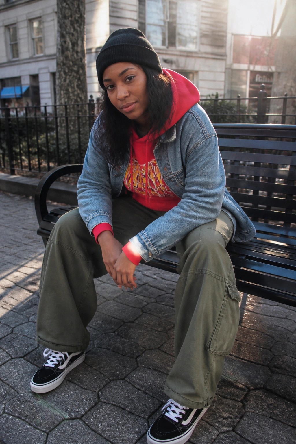 Woman in black beanie, red hoodie, denim jacket, and green cargo pants on bench