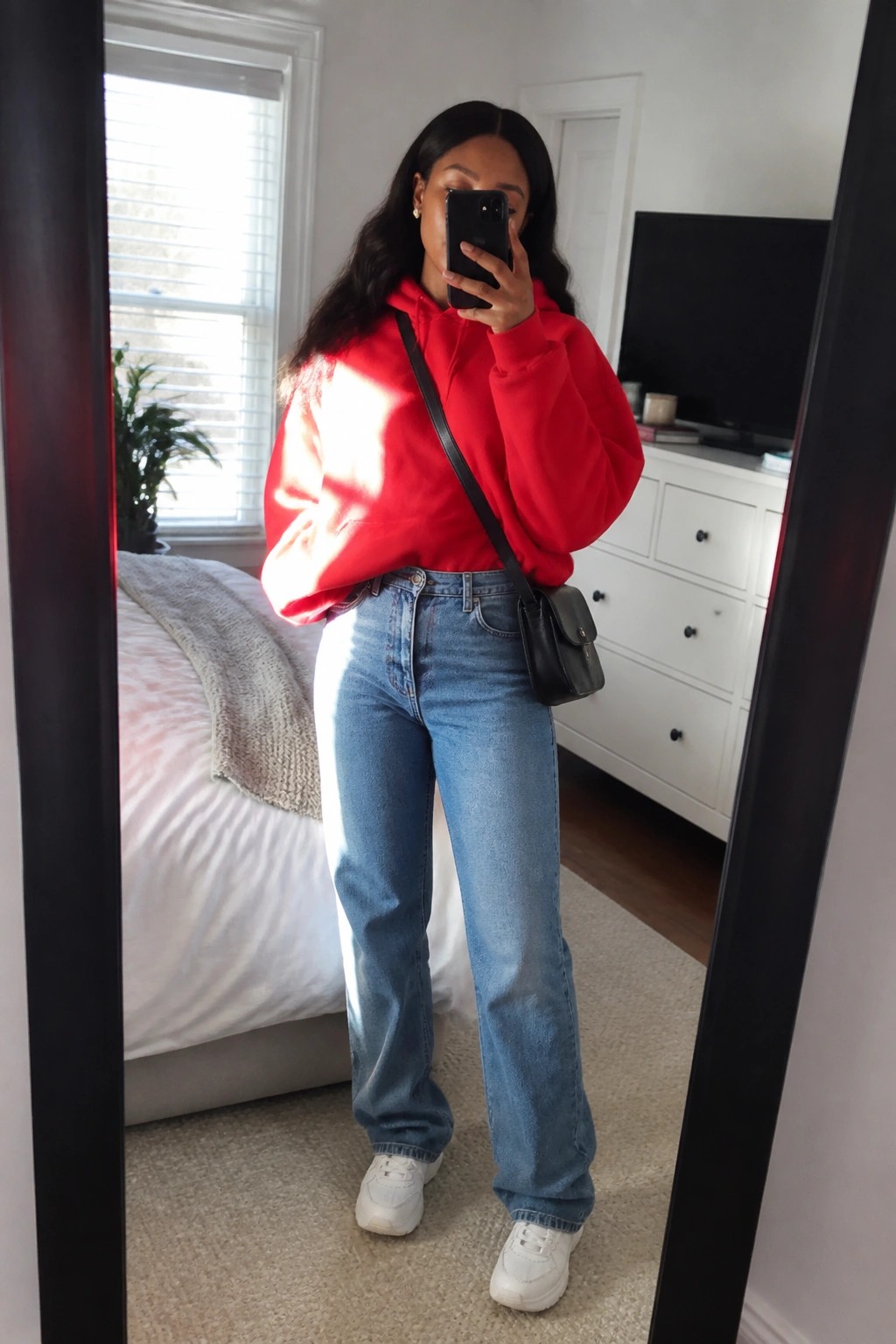 Woman wearing an oversized red hoodie tucked slightly into straight-leg blue jeans with white sneakers and a small black crossbody bag taking a mirror selfie in a bedroom