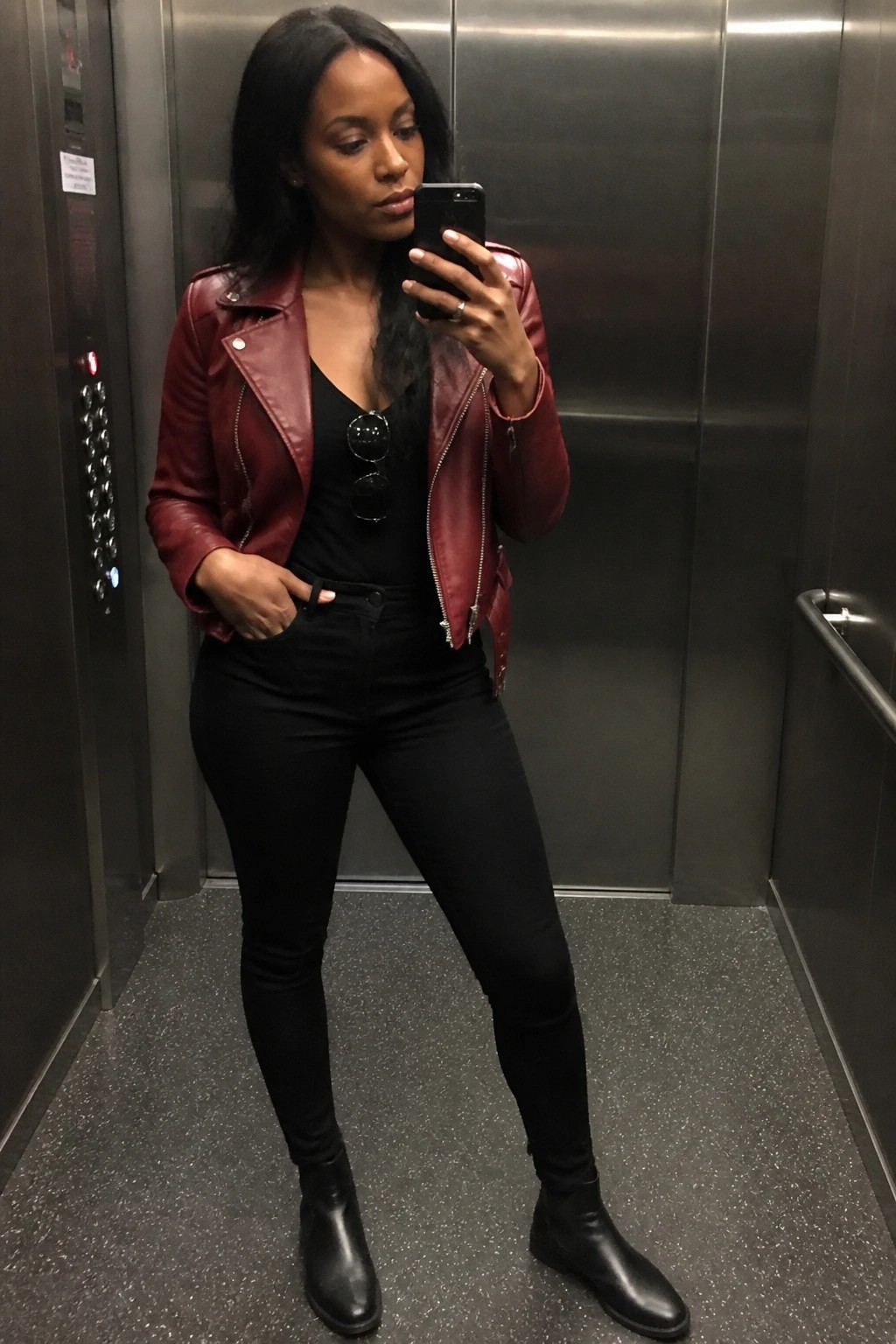 Woman in red leather jacket takes smartphone selfie inside elevator.