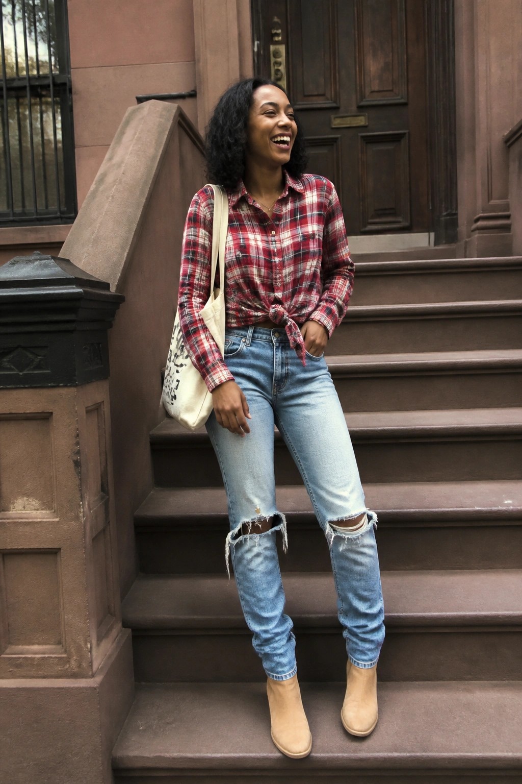 Woman in red plaid shirt and ripped jeans with tote bag on brownstone steps