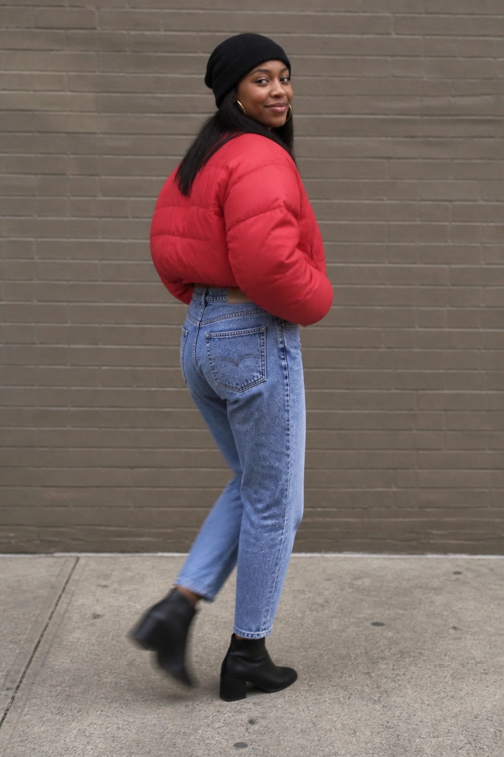 Woman in red puffer jacket, blue jeans, and black beanie on sidewalk by brick wall