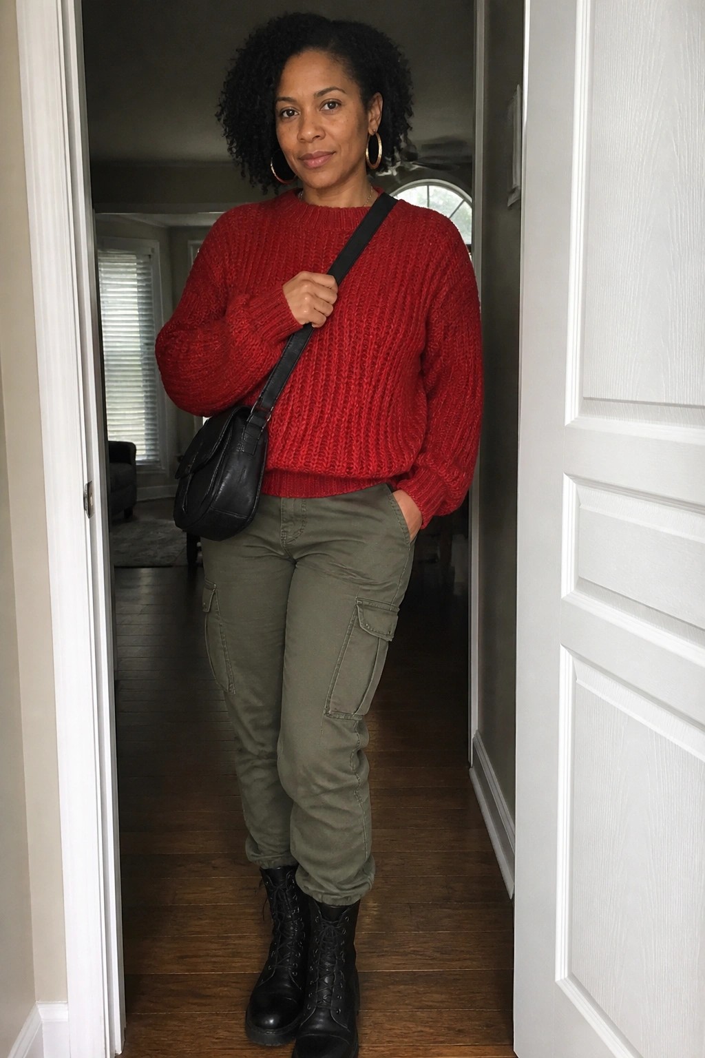 A Black woman in a red chunky knit sweater, olive cargo pants, black combat boots, gold hoop earrings, and a black crossbody bag standing in a doorway.