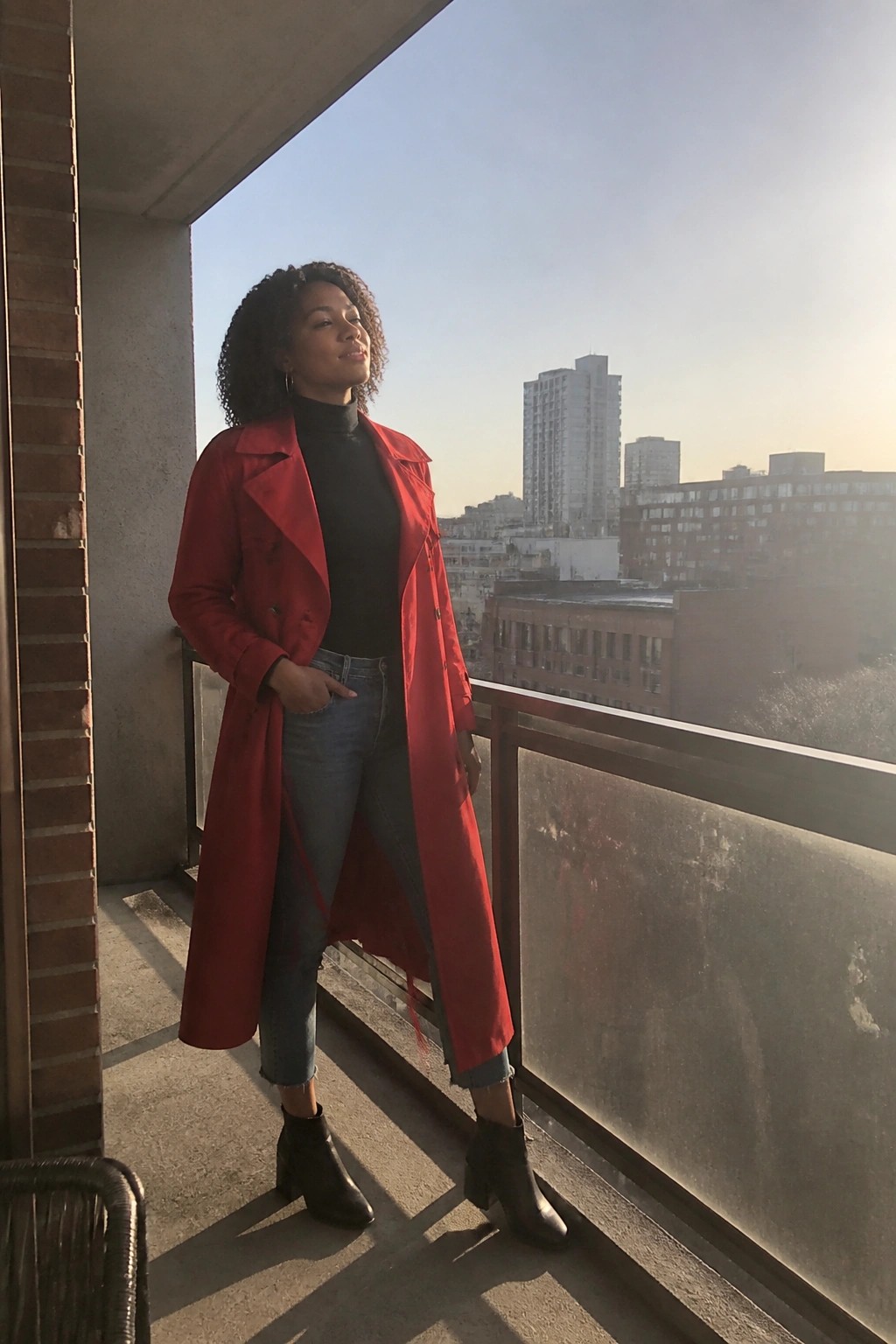 Woman in red trench coat on sunny balcony overlooking city skyline