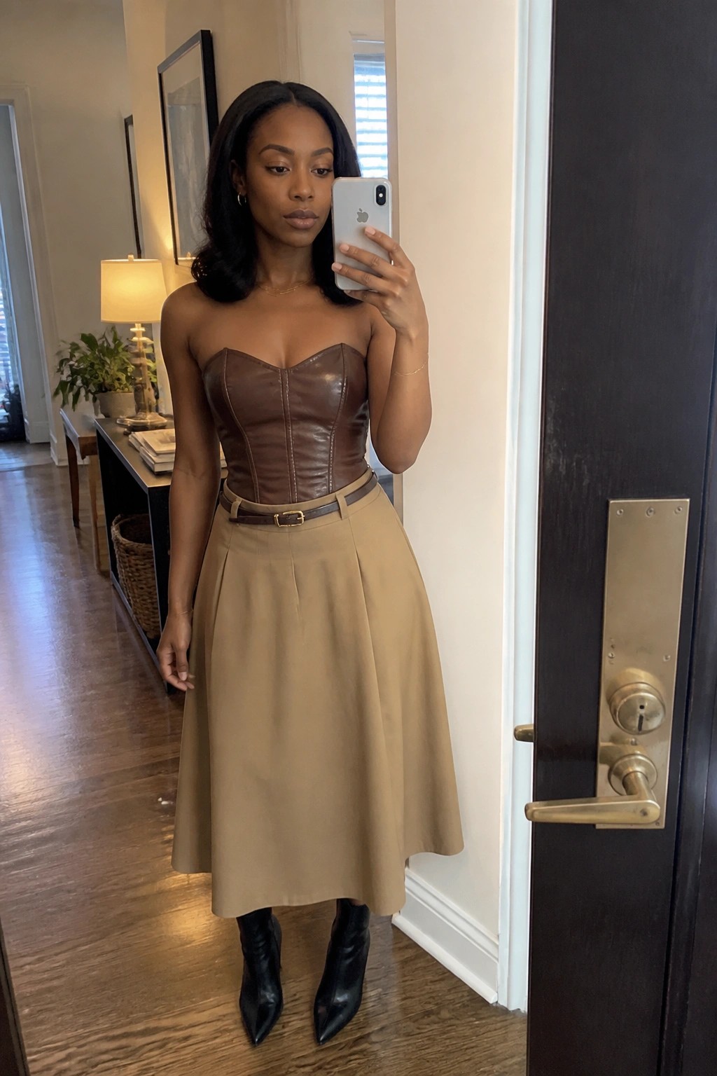 Woman in brown leather corset top and tan midi skirt taking mirror selfie