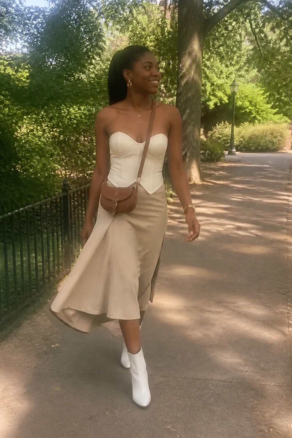 Woman in white corset top and beige skirt walks on sunny park path wearing white boots.