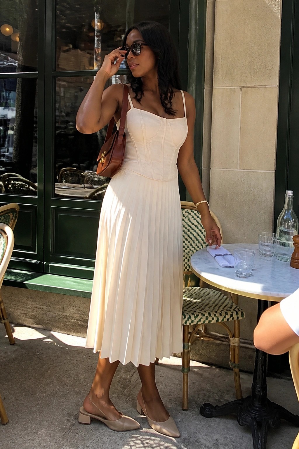 Woman in cream corset pleated dress adjusts sunglasses at outdoor cafe table.