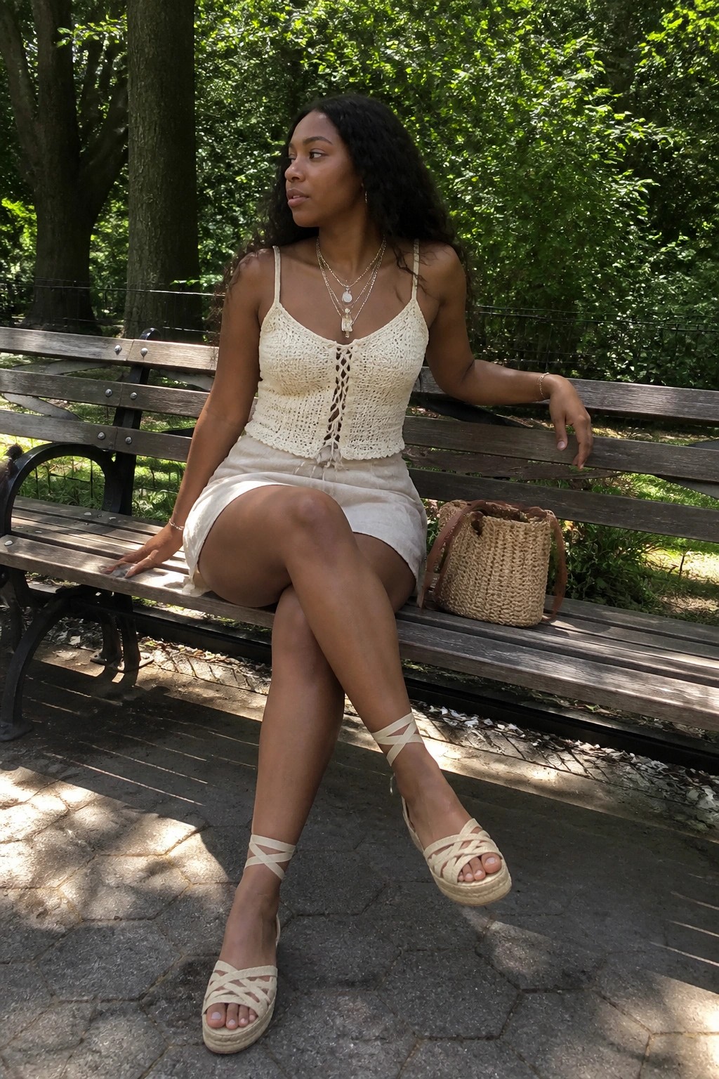 Woman in cream crochet top and skirt on park bench with straw bag.