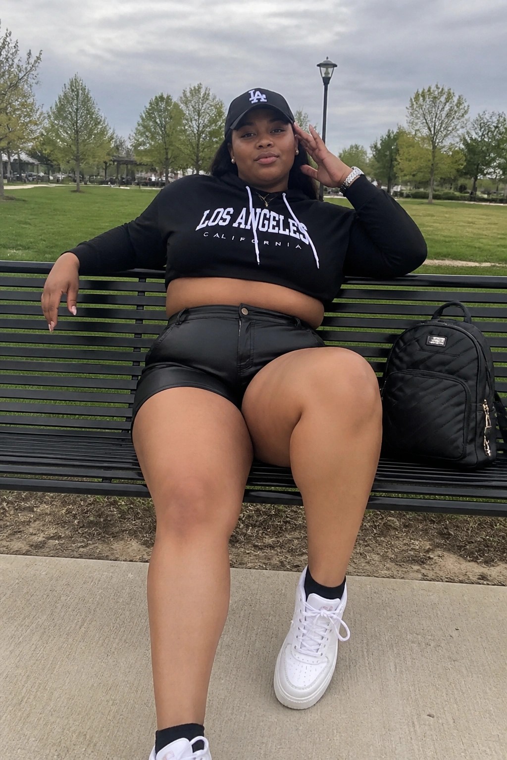 Plus size Black woman in black cropped Los Angeles hoodie with white text, black leather shorts, white sneakers, black LA cap, silver watch and necklace, seated on bench with black quilted backpack next to her.