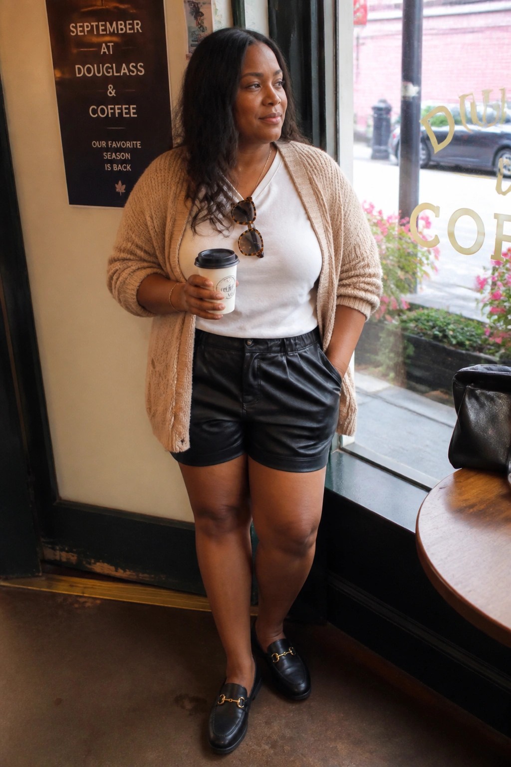 Plus size Black woman wearing black leather shorts, white crew neck tee, open beige knit cardigan, black loafers with gold hardware, tortoiseshell sunglasses tucked into neckline, holding a coffee cup inside a cafe.