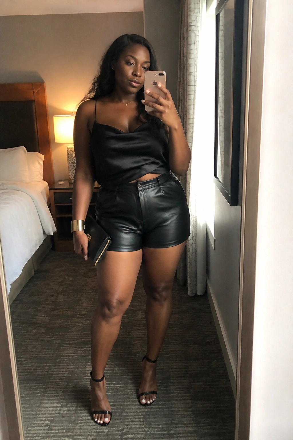 Woman in black satin camisole and leather shorts takes mirror selfie in hotel room.