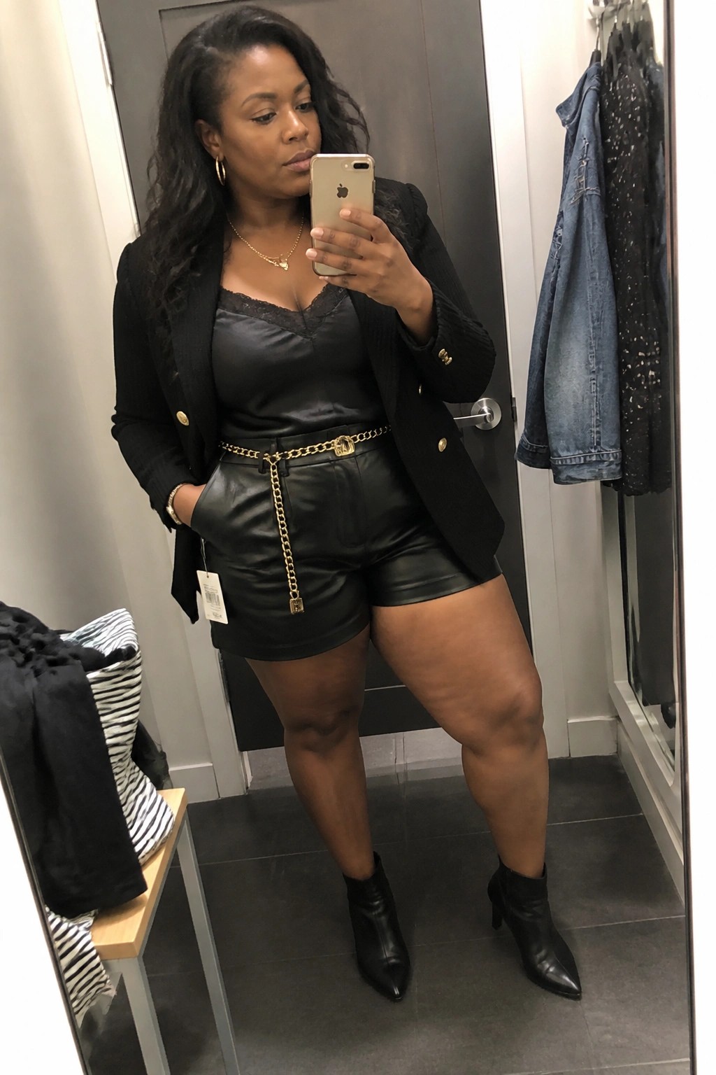 Woman in black blazer and leather shorts taking mirror selfie in fitting room