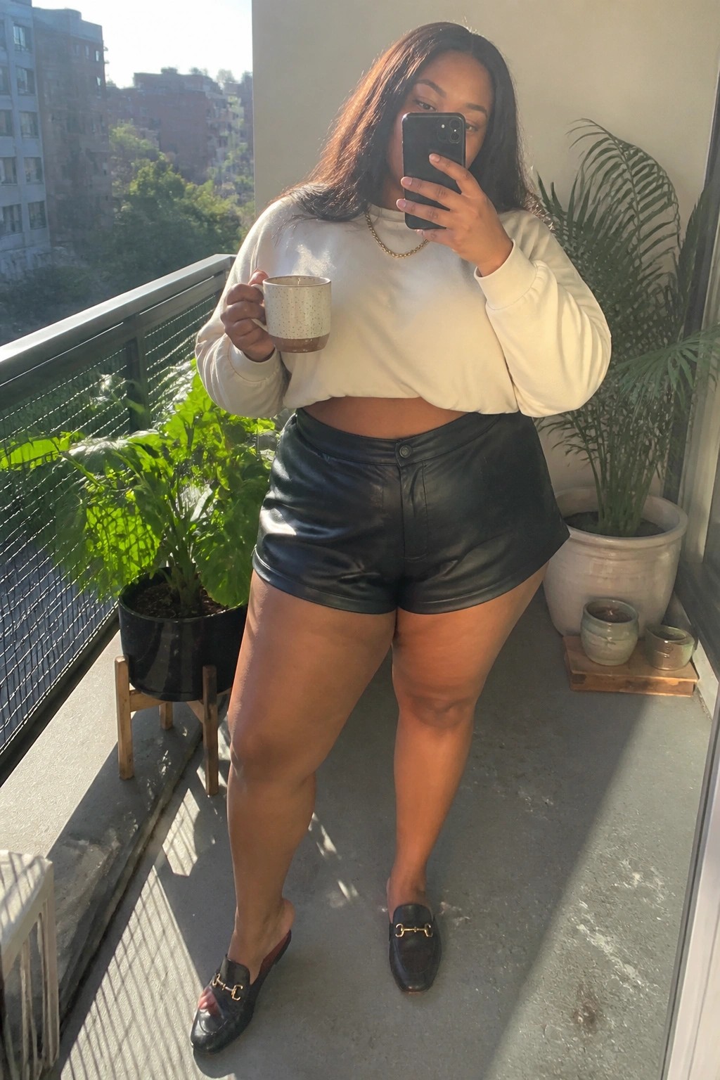 Woman in white cropped sweatshirt and black shorts taking selfie on sunny balcony with plants