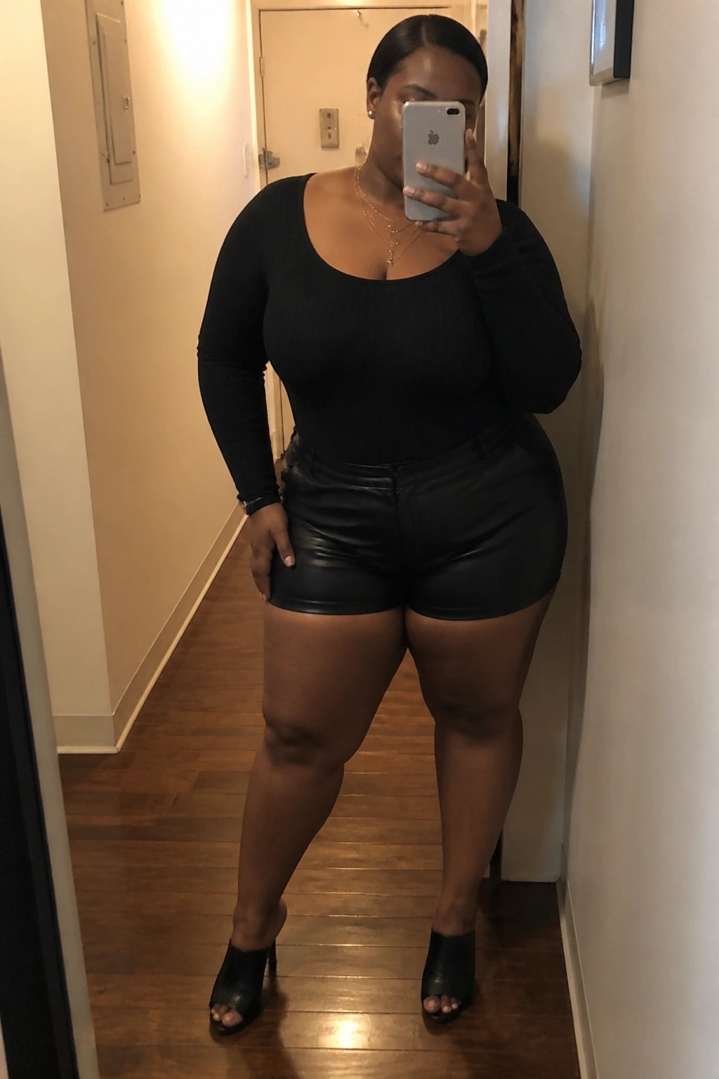 A plus size Black woman wearing black leather shorts, a black ribbed long sleeve scoop neck bodysuit, layered gold necklaces, and black open toe heeled mules while taking a mirror selfie in a hallway.